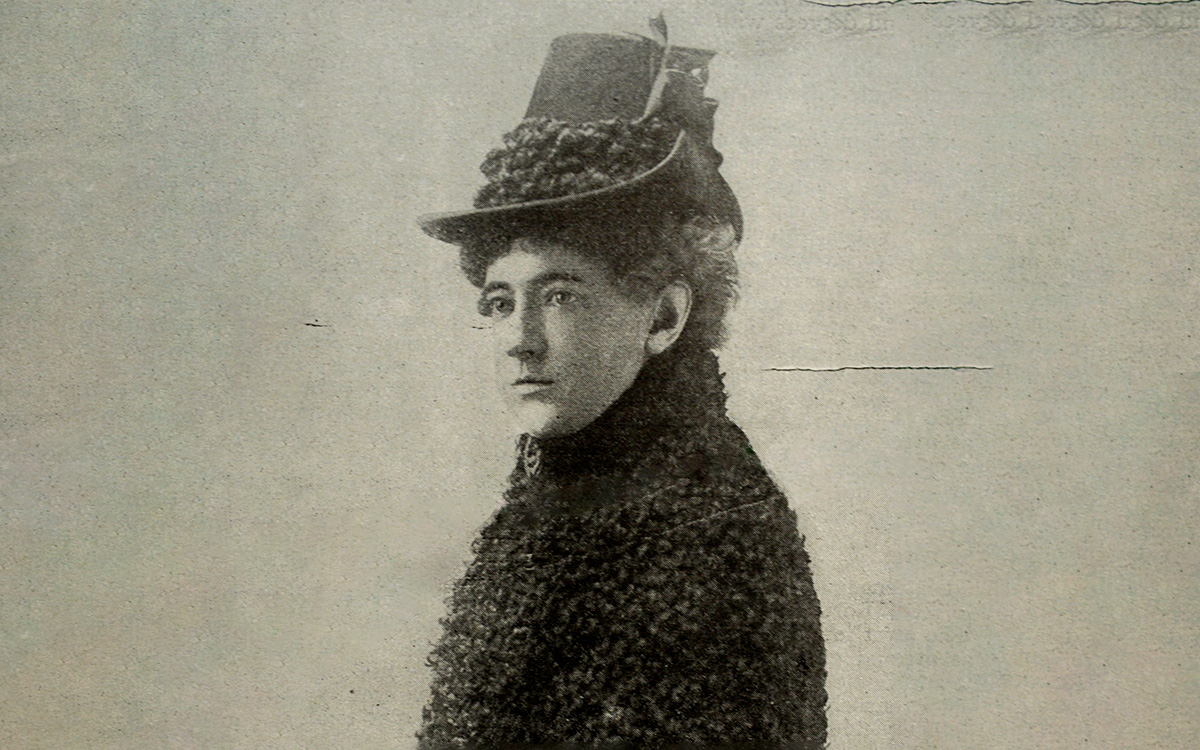 Image description: Black-and-white portrait photograph of Alice Sibley, wearing a dark-colored coat with a woolen texture and high collar, and a hat trimmed with a similarly-textured material.