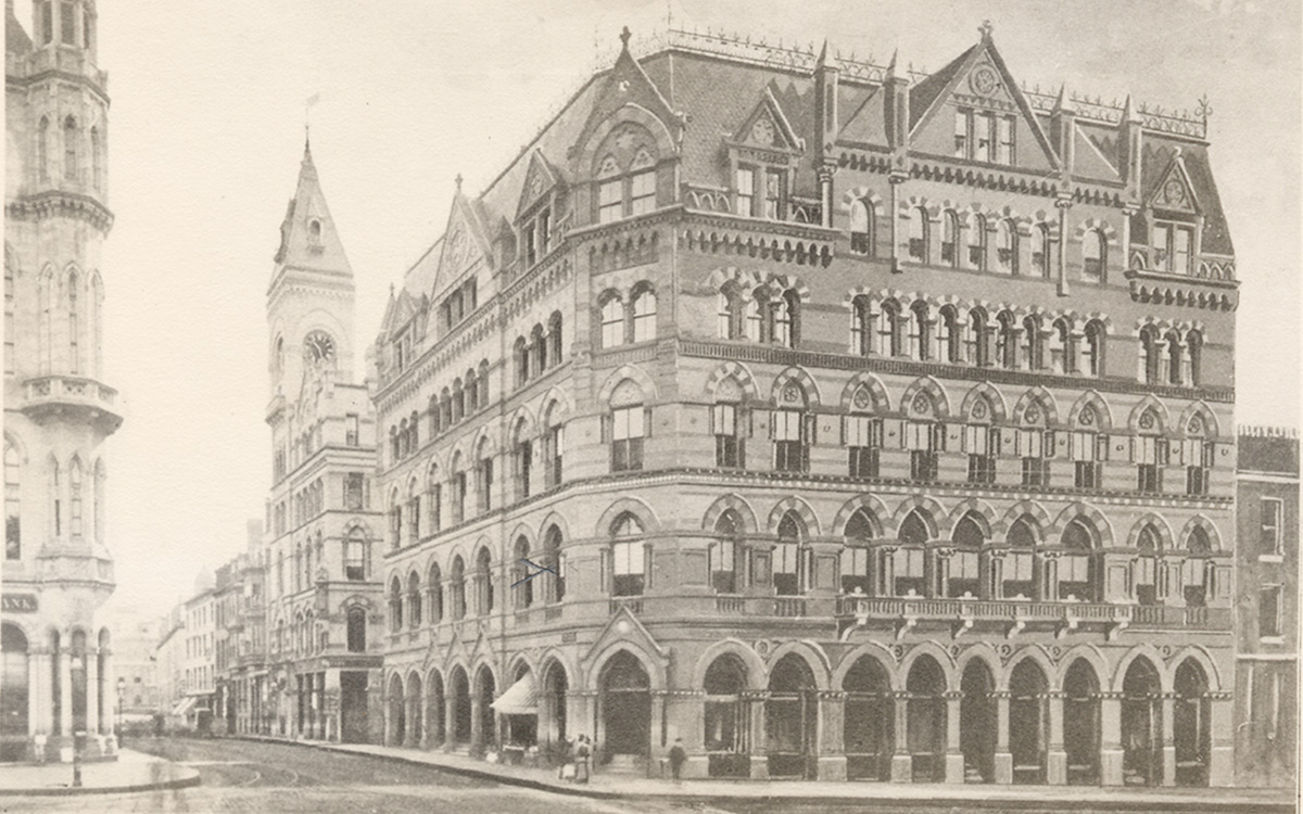 Image description: Sepia-toned photograph of the historic Hotel Boylston building in Boston, Massachusetts. Situated on a street corner, the building is six stories tall, with elaborate nineteeth-century architectural detail above the windows and on the roof. Further down the city street, other buildings, including a church edifice, are visible in the background.