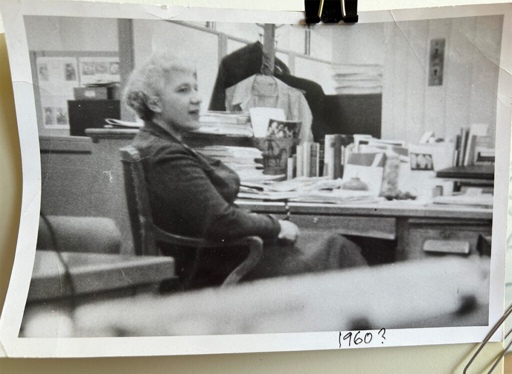 Candid photo taken from the side of a woman, Dorothy Adlow, sitting with a relaxed manner in a wooden desk chair at an office desk and talking to someone out of frame. The desk is close to the surrounding walls and is covered with books and papers. A coat rack can be seen in the background.