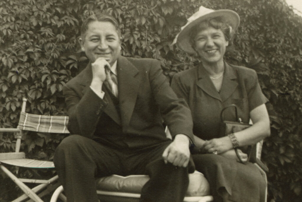 Smiling man (Nicholas Slonimsky) and woman (Dorothy Adlow), seated on a wide outdoor chair, with shrubs in the background. Slonimsky is dressed in a suit and rests his head casually on his hand. Adlow sits to his left, wearing a wide-brimmed hat and dress, with hands folded across the purse in her lap. 