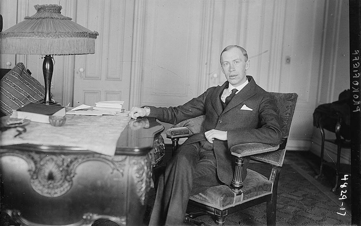 Black-and-white photo of Sergei Prokofiev, wearing a suit and tie and sitting at an embellished office desk. His posture is relaxed as he poses for the photo.