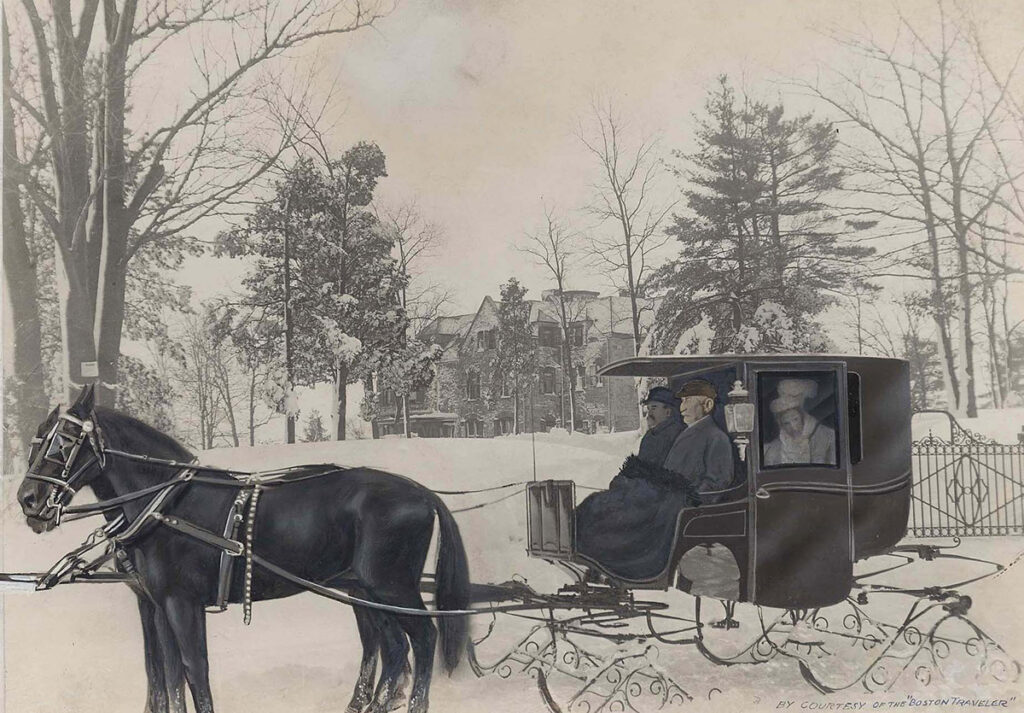 Image: A black-and-white photo of a horse-drawn enclosed carriage, with sleigh runners in place of wheels. Two black horses are harnessed to the carriage, and two men sit outside on the driver's bench of the carriage, bundled in coats. A woman (Mary Baker Eddy) sits inside the carriage, dressed in white and looking out the window. The carriage is on the snowy grounds of a large house, and a wide snow-covered lawn with trees around it separates the carriage and house.