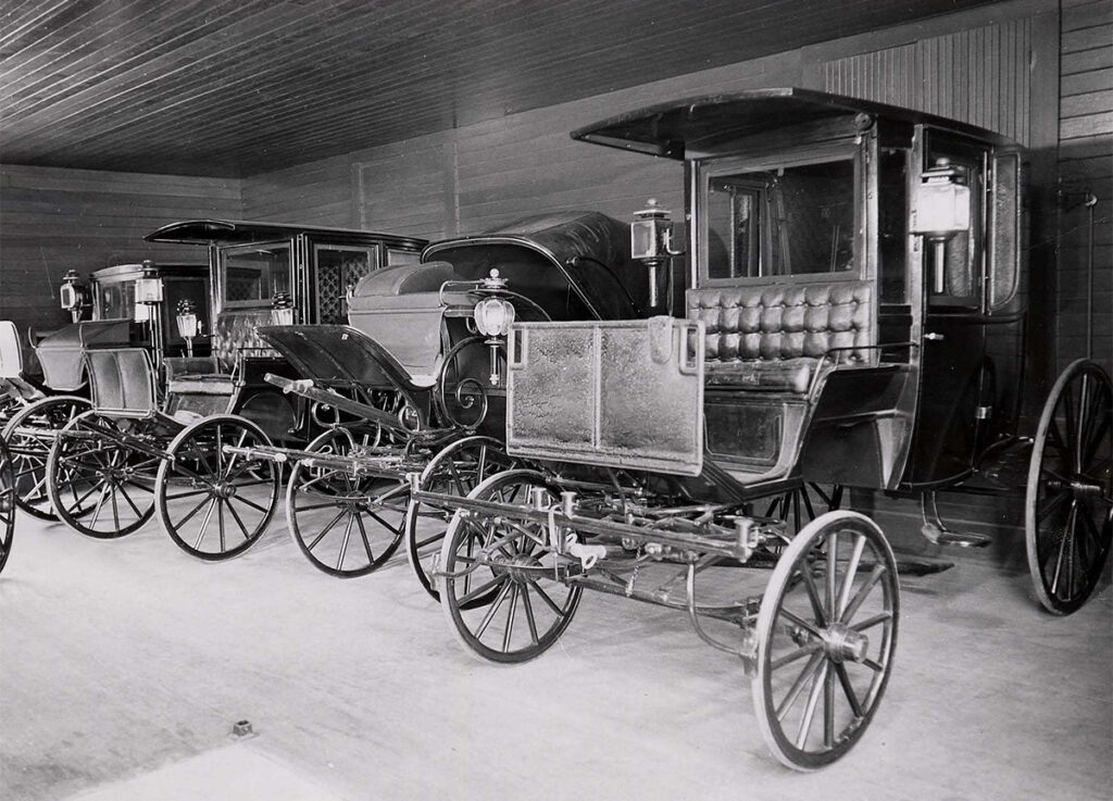 Image: A black-and-white photo of four 19th and 20th century-era carriages of varying styles, parked in an interior garage/shed. The wheel of a fifth carriage is visible at the left edge of the photo.