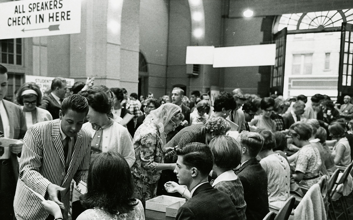 2025-02a Biennial meetings - F284880_1960s_Photo_RegistrationTable_001 FINAL Image description: A black-and-white photograph showing the inside of a well-lit entrance hall with light-colored stone walls. The room is full of people, most of whom appear to be in their teens or twenties. Several individuals sit along a long table being used for registration, while queues of people wait to talk to those at the table. A sign hanging from the ceiling reads "All Speakers Check In Here," with an arrow pointing to another part of the room. The scene is lively and bustling.