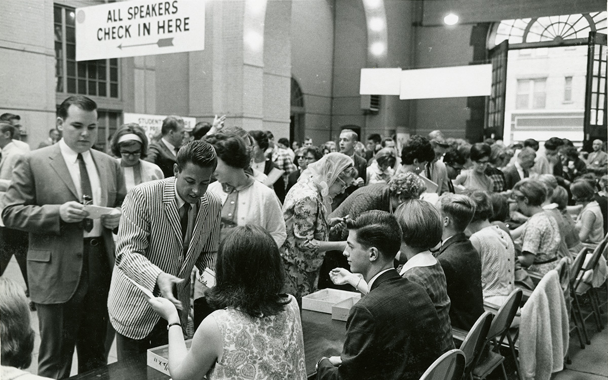 Image description: A black-and-white photograph showing the inside of a well-lit entrance hall with light-colored stone walls. The room is full of people, most of whom appear to be in their teens or twenties. Several individuals sit along a long table being used for registration, while queues of people wait to talk to those at the table. A sign hanging from the ceiling reads "All Speakers Check In Here," with an arrow pointing to another part of the room. The scene is lively and bustling.