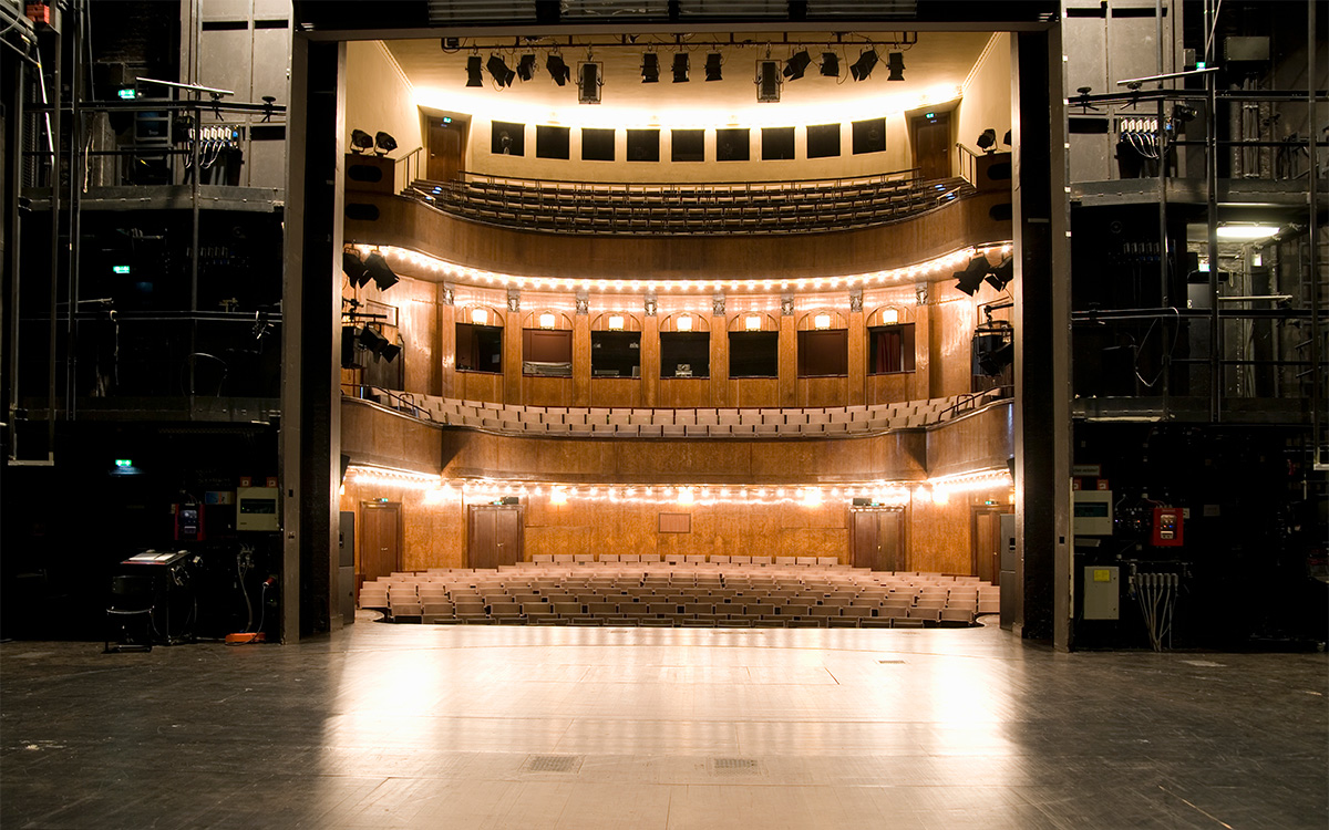 Image description: Photo of a stage in a multi-level theater, from the perspective of someone standing on the stage and looking towards the auditorium. The auditorium is empty and lit, and the lights are reflecting back onto the stage. Part of the backstage hardware (curtains, scaffolding, etc.) can be seen to the left and right of the stage itself.