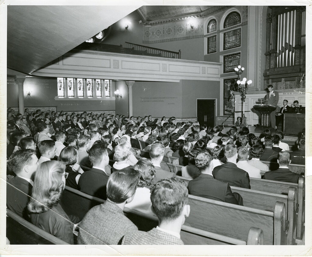 Image description: A black-and-white photograph of the inside of a church (the Original Edifice of The Mother Church), with the pews full of young adults in suit jackets and tidy church clothing. A man in a suit stands at the speaker's podium at the front of the room.