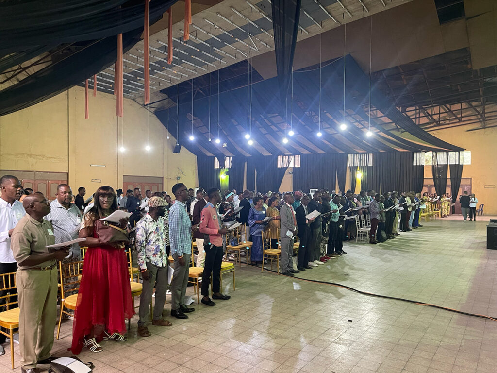 Image description: Photo of a large conference space or church hall full of people, many in formal dress (such as suit jackets or dresses) and others in business-casual attire. They are standing and singing in rows, each in front of their chairs, which are set up in many single rows for attendance at a presentation.