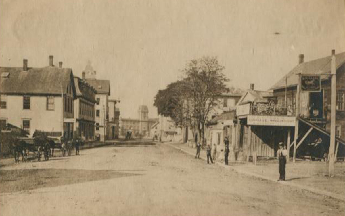 Image description: Sepia-toned photo of a small-town street in the late 1800s. The road is unpaved, with carriages seen on the left.