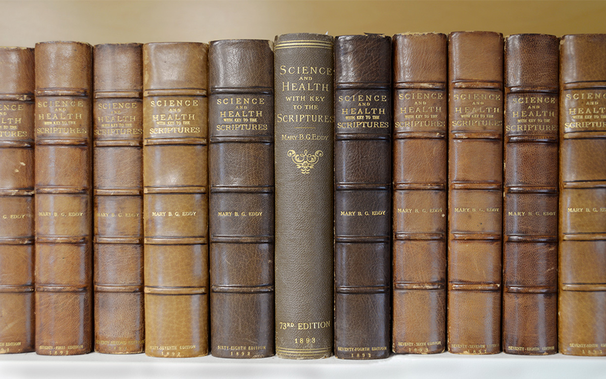 Image description: A row of about 10 brown books on a bookshelf, editions of Science and Health with Key to the Scriptures. The spine of the book in the center reads "Science and Health with Key to the Scriptures, Mary B. G. Eddy, 73rd Edition, 1893."