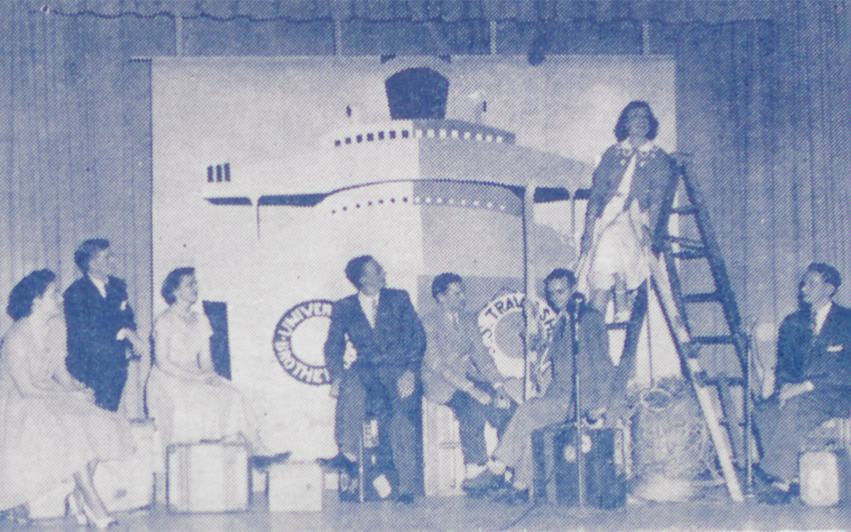 Image description: Black-and-white photo showing several young men and women, dressed in the fashion of the 1950s, on a stage. Suitcases are set up as props, on which the people are seated, and one person is seated partway up a ladder. A backdrop showing a large ship hangs behind them, and a microphone is at the front of the stage on a mic stand.