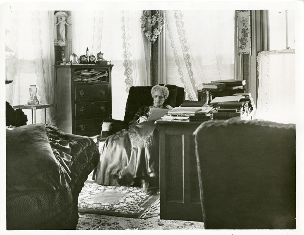 Image description: Black and white photograph of Mary Baker Eddy, sitting at her desk in an office with a large bay window with gauzy curtains. The desk is piled high with books, and a tall cabinet is on the other side of her. She is holding a piece of paper and a pen. 