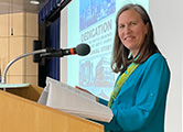 Cindy at podium at BSH LA - CROP Image description: Photograph of Cindy Safronoff at a podium, next to a large screen, giving a presentation.
