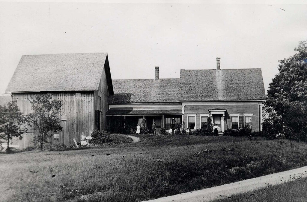 Image description: Black and white photograph of a farmhouse and attached barn, with open sky in the background, a sizeable lawn in front, and some young trees lining one edge of the house. Although small in the photo and difficult to see (due to the distance of the photographer), members of a family pose outside the house, lined up in a row.