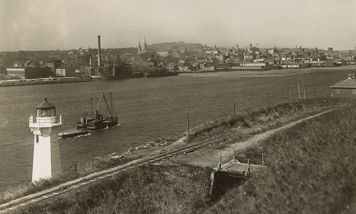 Image description: Sepia-toned photograph shows a channel of water, looking across the water to buildings and city landmarks on the opposite shore. A small white lighthouse is at the bottom left of the image, and a vessel sails up the channel.