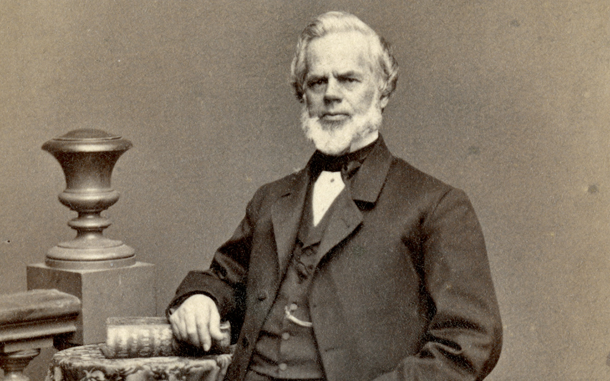 Image description: Antiquated black-and-white photograph of Phineas P. Quimby, seated and posing for a portrait. He is seated, wearing a three-piece suit and bow tie, and is resting his arm on a large book placed on a side table next to him. To his right is a fixture resembling a staircase bannister.