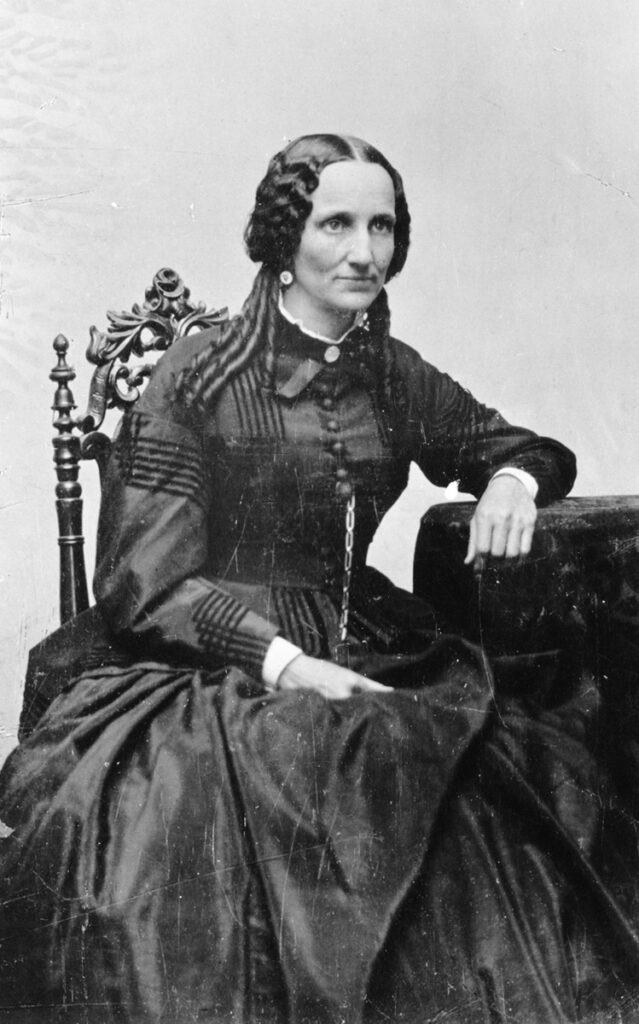 Image description: Black-and-white photo of Mary Baker Eddy, posed for a portrait, seated in a chair with an ornately-carved back and resting her hand on a table next to her. She is wearing a dark-colored long-sleeve dress with striped sleeve detail and a long row of buttons, and she is looking off to the left of the camera.
