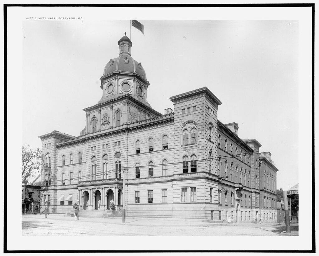 Image description: Black-and-white photograph of a large four-story municipal building, with a dome complete with cupola and an American flag flying from the top.