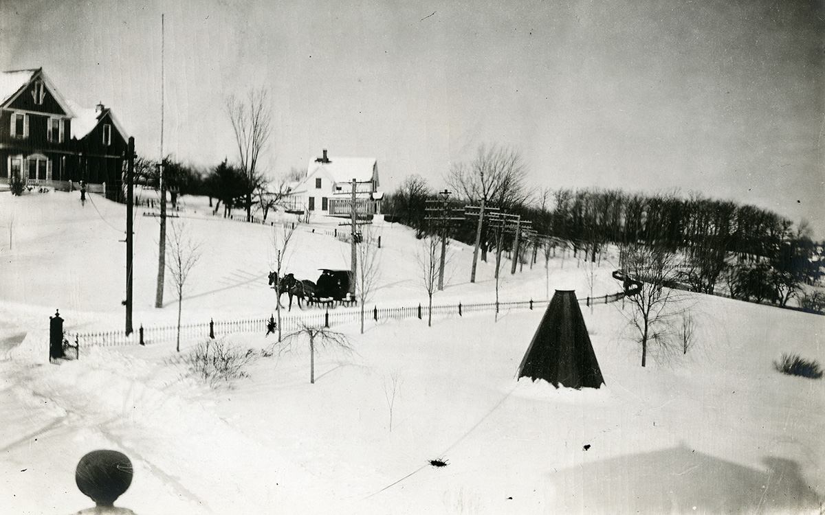 Image description: Black and white photograph of a snowy New England country scene. A country road cuts across the middle of the photo, on which a carriage is pulled by two horses. On the other side of the road, in the background, two large houses sit on a hill with a fence in between their properties. The hillside is sparsely wooded, with a denser forest in the distance.