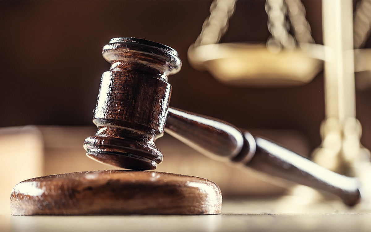 Image description: Still life photograph of a dark wooden gavel resting on a round wood block on a desktop. In the background, blurred, is a set of old-fashioned looking gold metallic scales, representing the scales of justice.