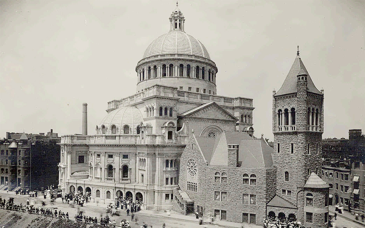 Image description: Black-and-white photograph of The Mother Church, at an wide angle that shows both the original edifice and the larger Extension next to it. Groups of people can be seen entering and exiting the two buildings.