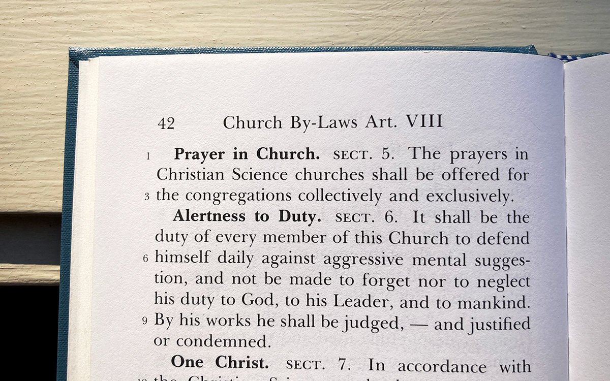 Image description: Color photograph of a page from the Manual of The Mother Church, showing the top left corner of the book with the "Prayer in Church" By-law visible on the page. The book is resting on an off-white piece of painted wood.